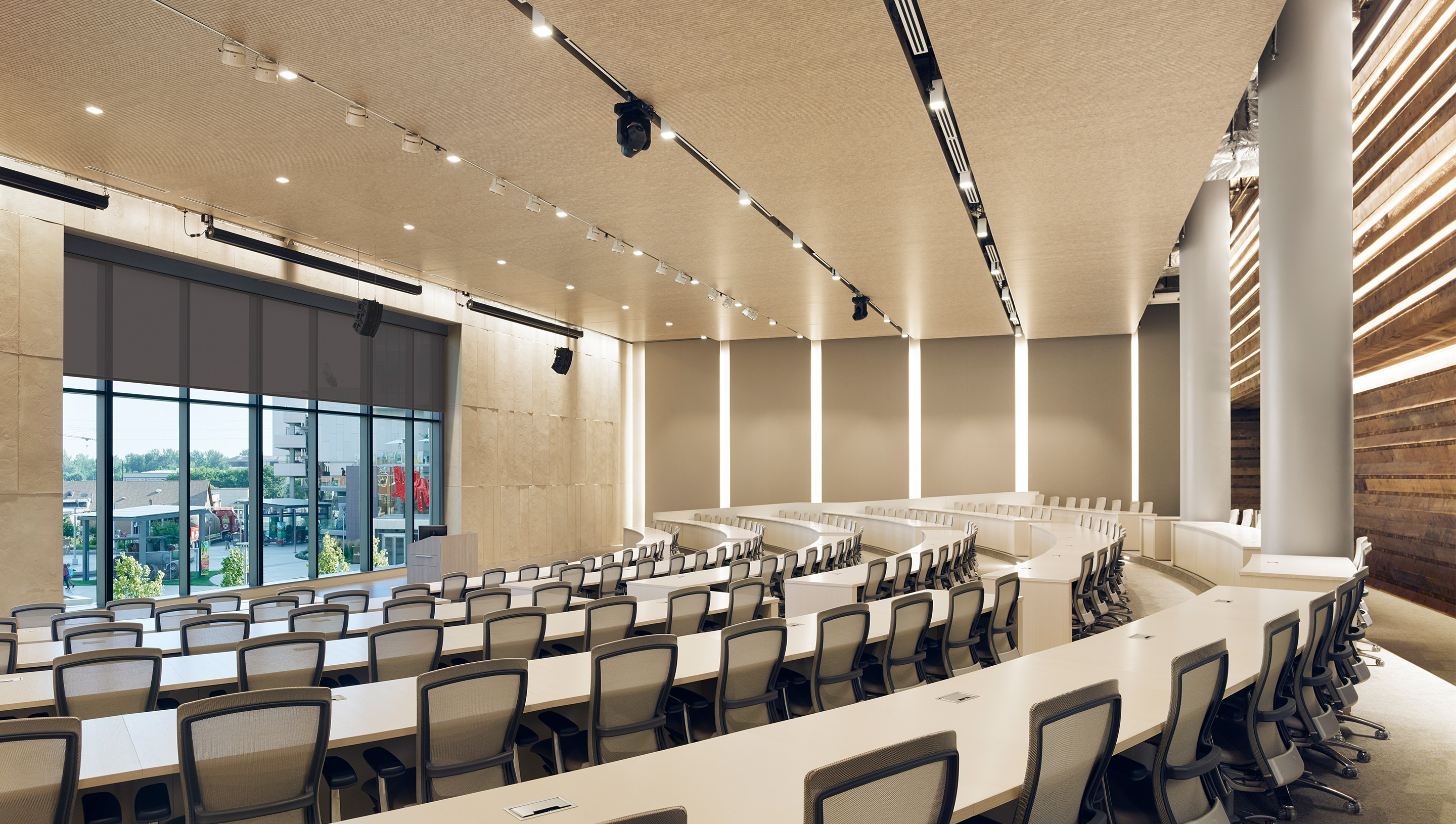 Auditorium with empty seats, lectern, windows with dark colored roller shades, and linear LED lighting integrated into the wall panels.