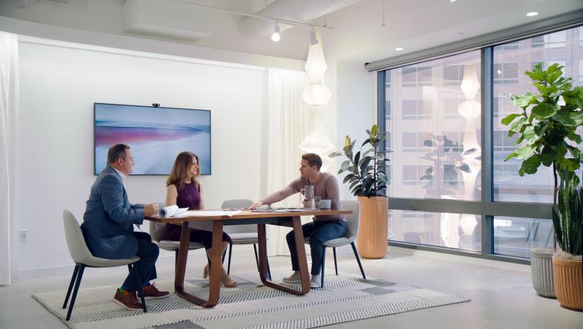 Three people having a meeting at a table in a room with potted plants and evocative light fixtures.
