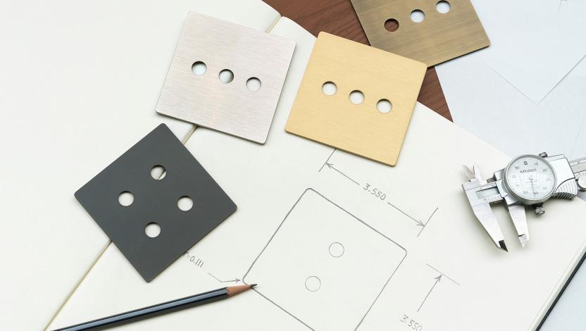 Working surface with sketch of a faceplate, drawing pencil, and several of the Signature Metal faceplates in different finishes.