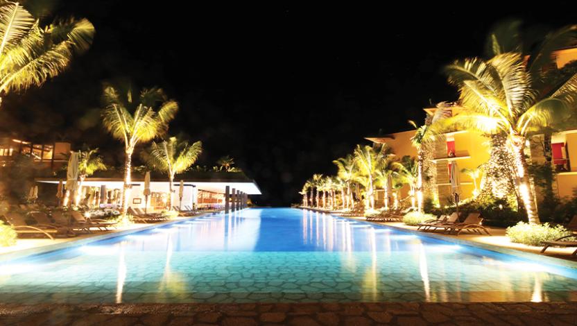 A nighttime view of Hotel Xcaret Mexico that includes blue pool and uplit tropical trees. 