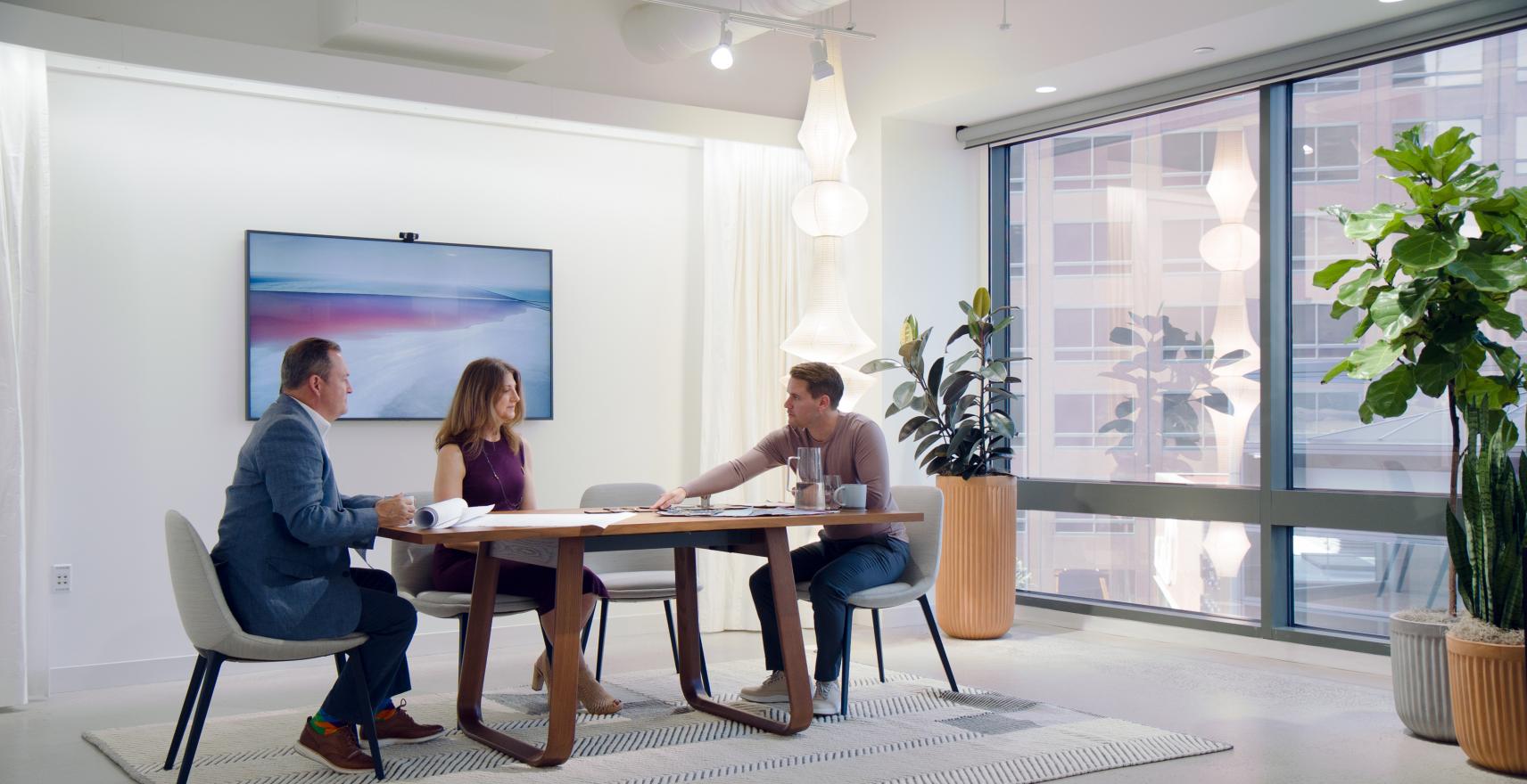 Three people having a meeting at a table in a room with potted plants and evocative light fixtures.