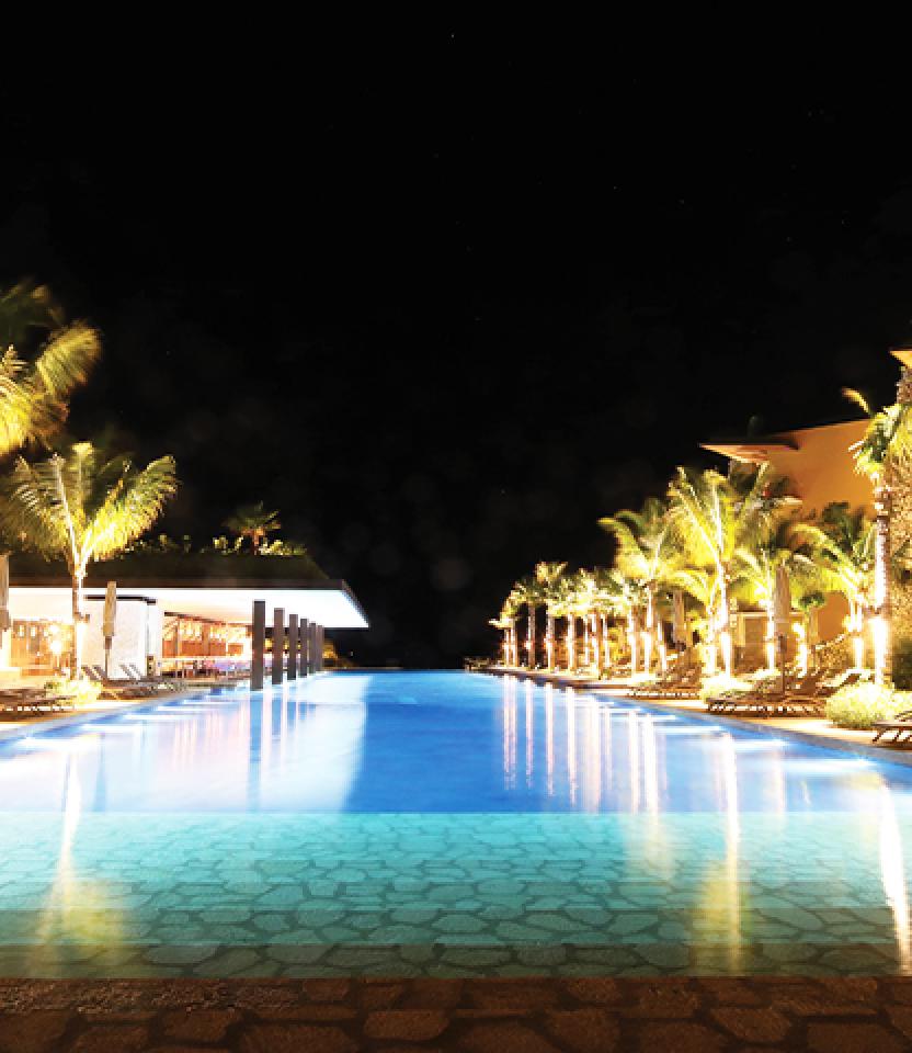 A nighttime view of Hotel Xcaret Mexico that includes blue pool and uplit tropical trees. 