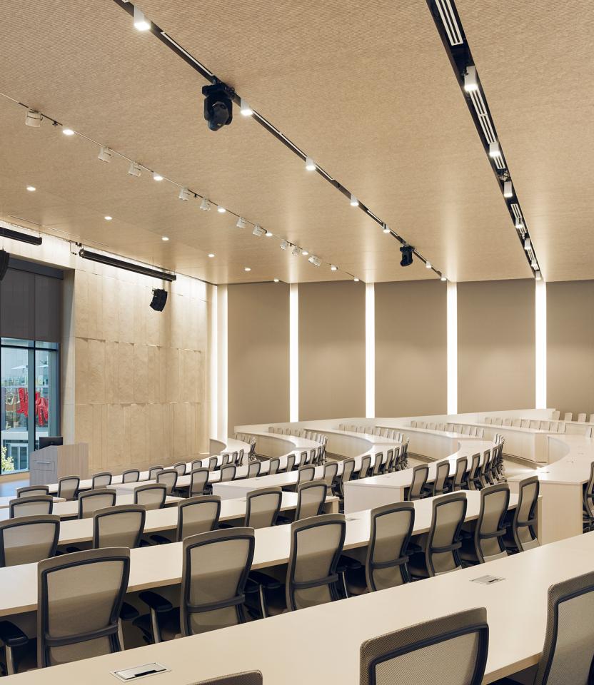 Auditorium with empty seats, lectern, windows with dark colored roller shades, and linear LED lighting integrated into the wall panels.