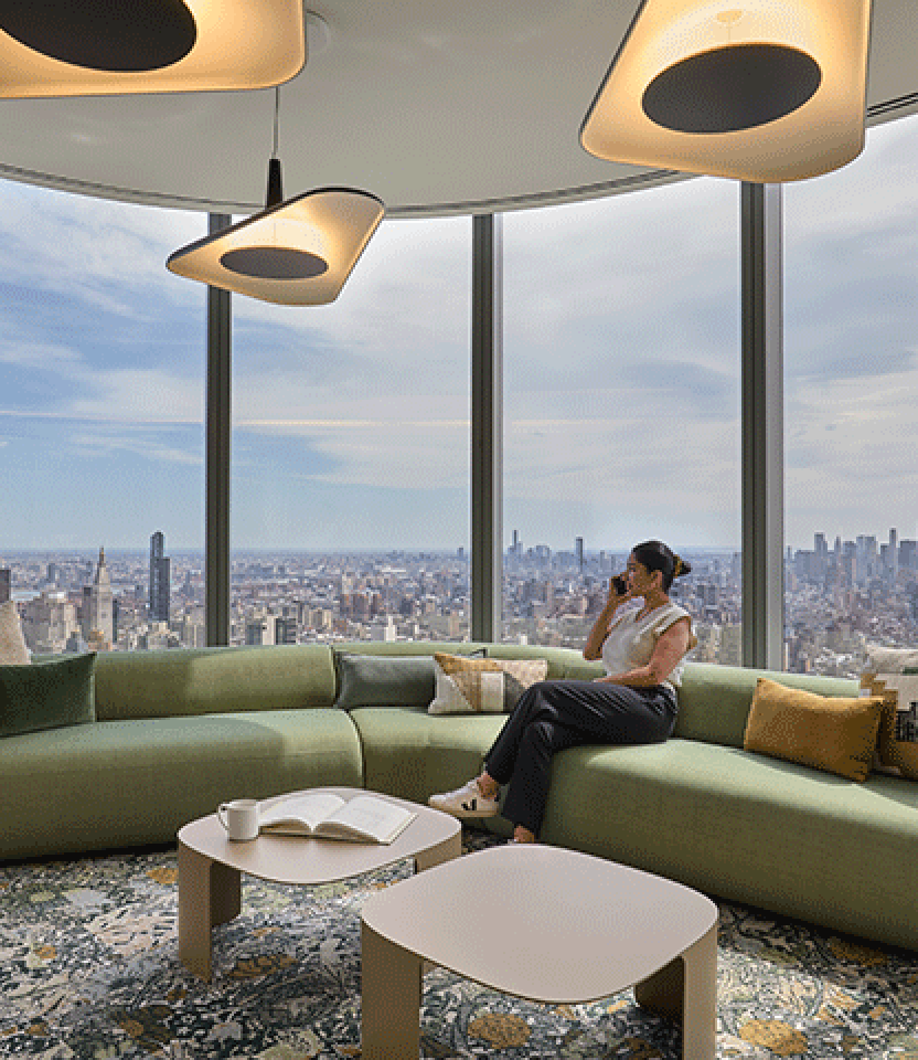 A woman sits on a low green sofa in a modern office. The skyline of New York is visible out the wall of windows