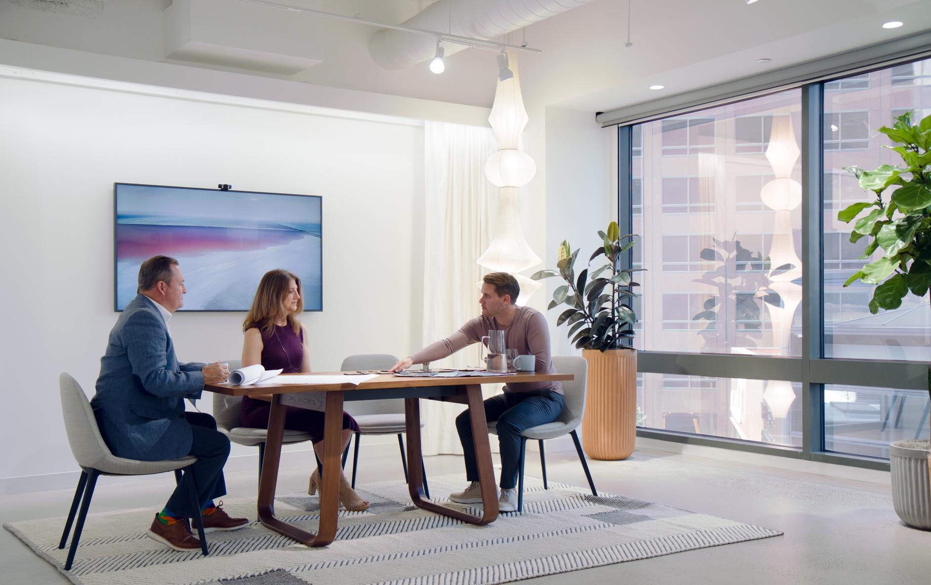 Three people having a meeting at a table in a room with potted plants and evocative light fixtures.