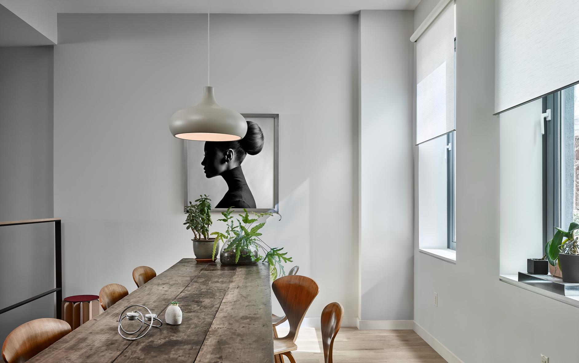 Stylish dining area with wood dining table, black-and-white photograph of a woman’s profile, and two windows with white roller shades.