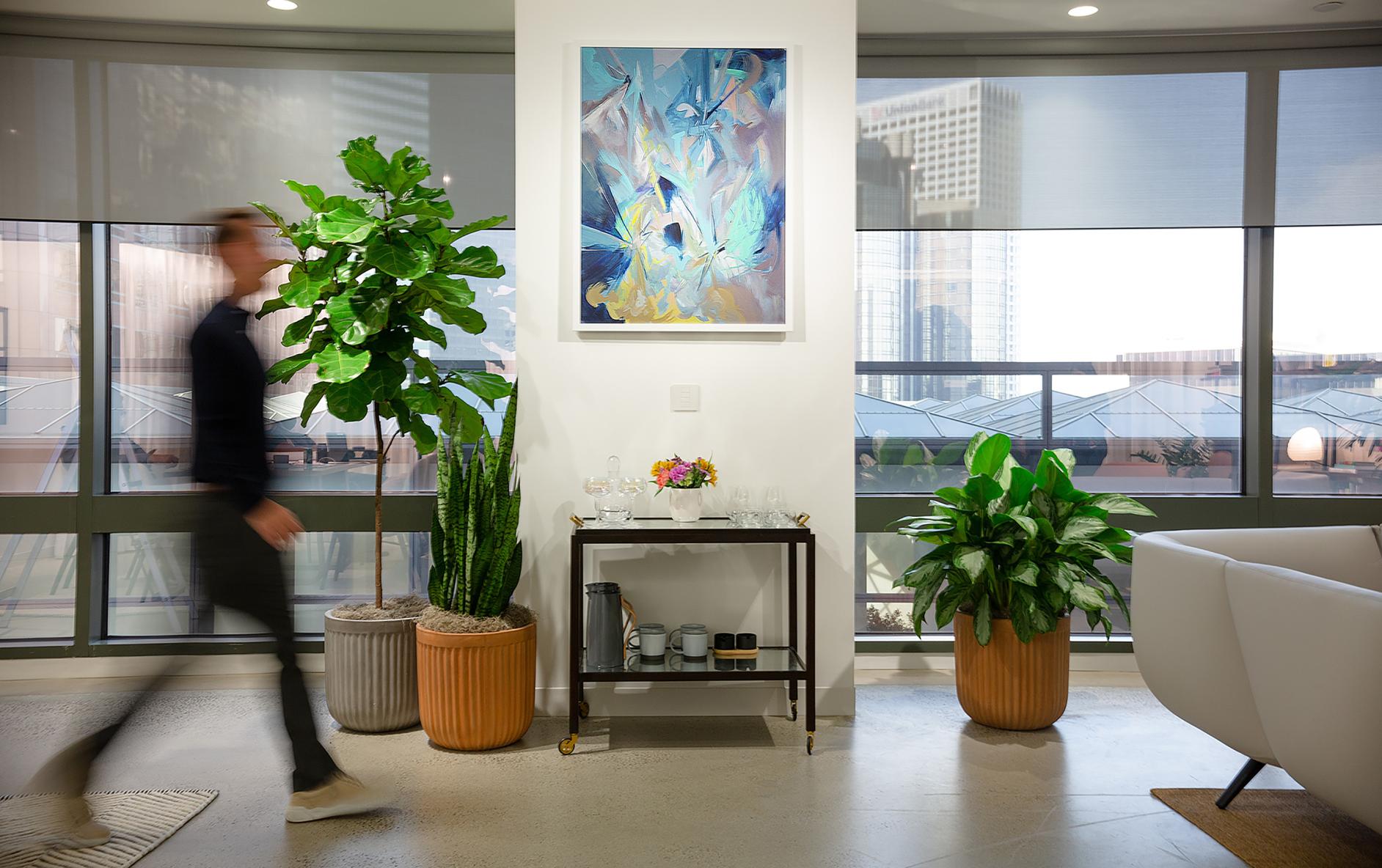 Contemporary office interior with neutral palette, tile floor, several potted plants, and grey roller fabric on the windows. 